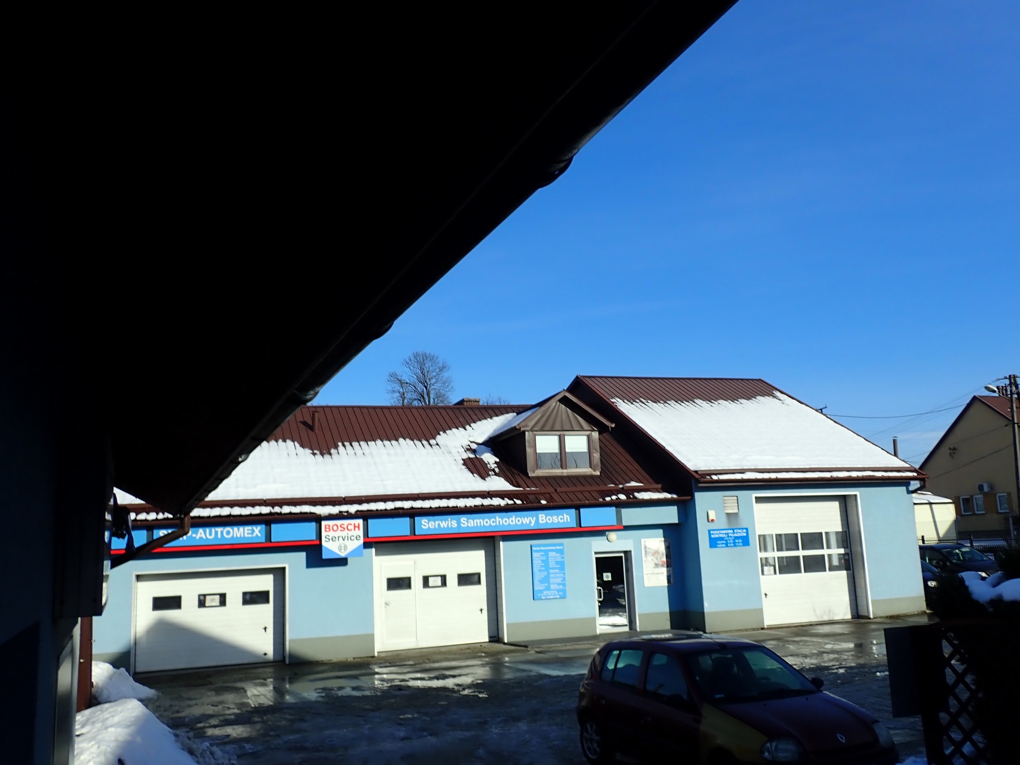 Zimowy widok na budynek serwisu samochodowego Bosch z trzema bramami garażowymi i zaparkowanym czerwonym samochodem osobowym na ośnieżonym placu.