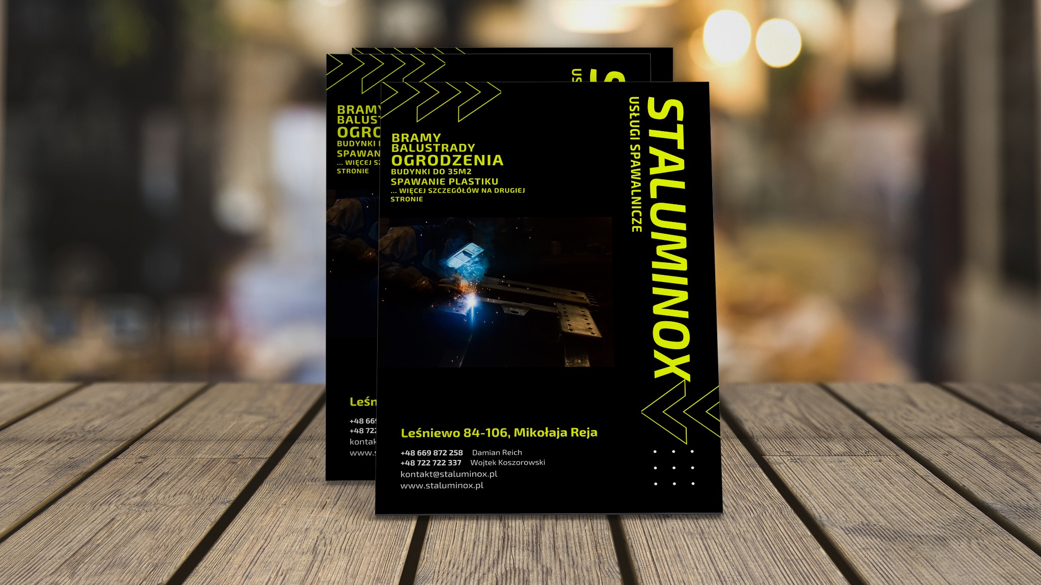 Ulotki reklamowe firmy spawalniczej Staluminox na drewnianym stole, prezentujące usługi spawania bram, balustrad i ogrodzeń oraz spawanie plastiku.