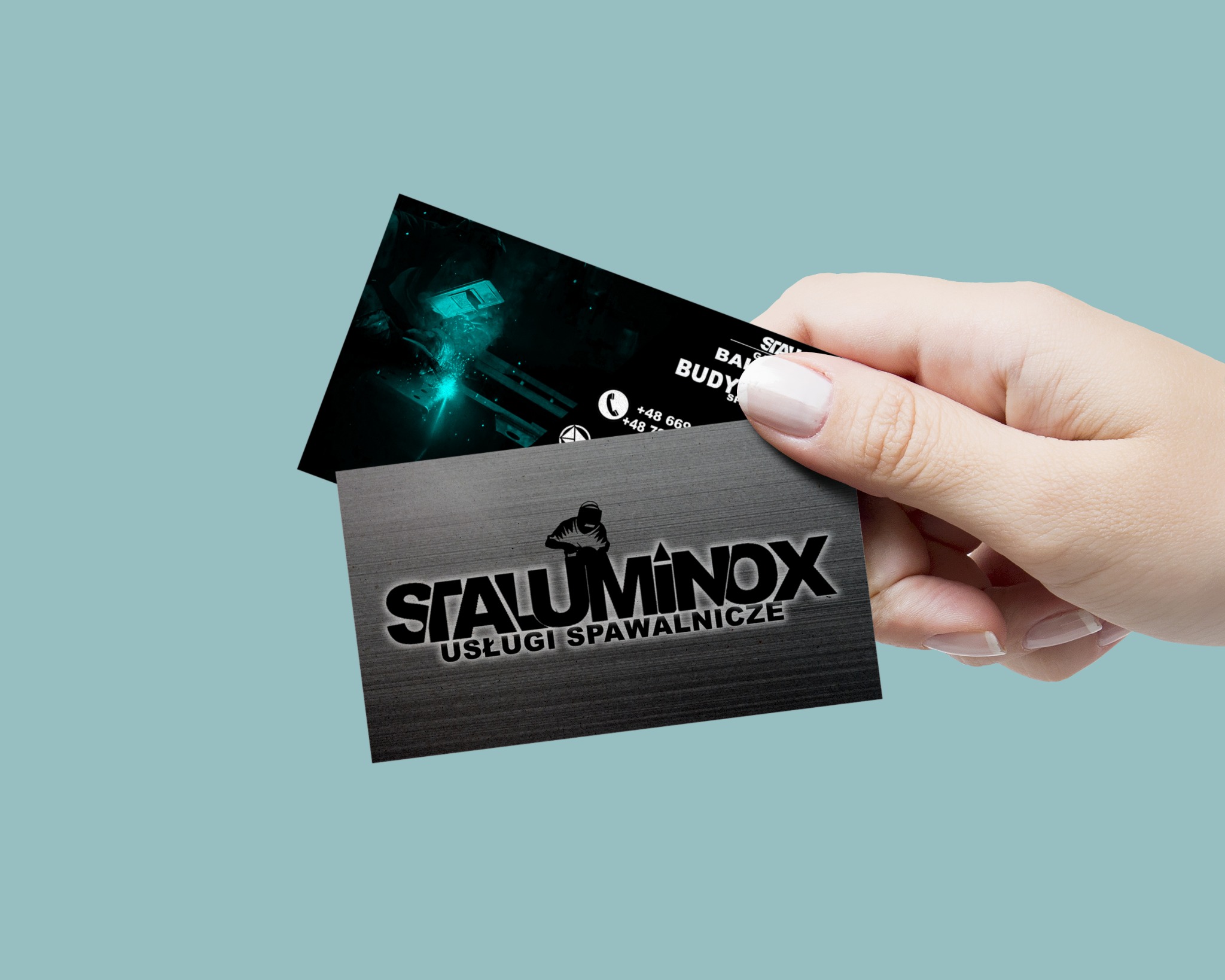 Dłoń trzymająca dwie wizytówki firmy Staluminox oferującej usługi spawalnicze. Jedna wizytówka ukazuje spawacza podczas pracy w niebieskiej tonacji, druga ma metaliczne wykończenie z logo firmy.