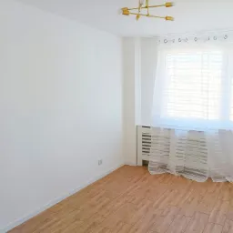 Pusty pok&oacute;j z gładkimi, białymi ścianami, panelami podłogowymi imitującymi drewno, grzejnikiem pod oknem z delikatną, prześwitującą firanką i nowoczesną, złotą lampą sufitową.