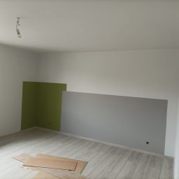 KOLORGIPS - remonty i wykończenia wnętrz - Świeżo pomalowane ściany w pokoju, fragmenty w kolorze zielonym i szarym, podłoga wyłożona panelami. Na podłodze leżą kartony zabezpieczające.