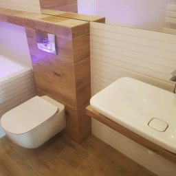 KOLORGIPS - remonty i wykończenia wnętrz - Nowoczesna łazienka z podwieszaną toaletą, umywalką na drewnianym blacie i płytkami imitującymi drewno. Minimalistyczny design, subtelne oświetlenie.