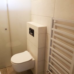 KOLORGIPS - remonty i wykończenia wnętrz - Nowoczesna łazienka z podwieszaną toaletą, grzejnikiem drabinkowym i kabiną prysznicową. Jasne płytki imitujące kamień. Minimalistyczny design.