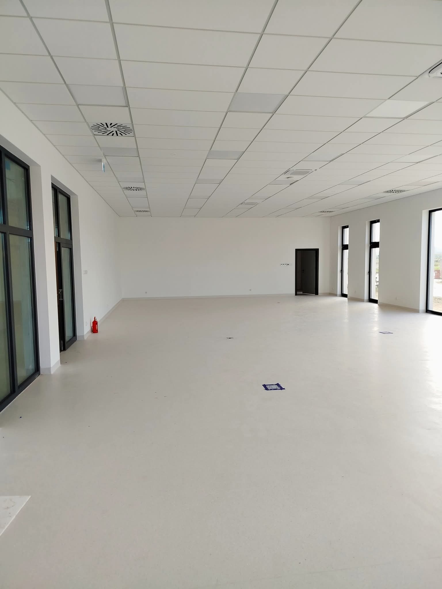 Jasne, przestronne wnętrze po remoncie, z białymi ścianami, podwieszanym sufitem z kratką i dużymi oknami. Widoczny fragment gaśnicy przy ścianie.