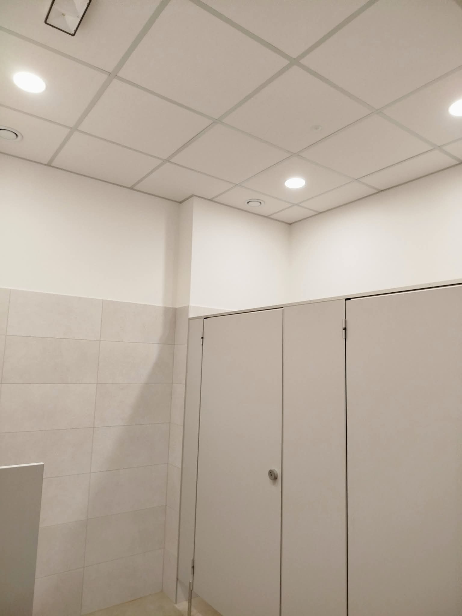 Wnętrze wykończonej toalety publicznej z kabinami, jasnymi płytkami na ścianach i podwieszanym sufitem z oświetleniem punktowym. Minimalistyczny design.