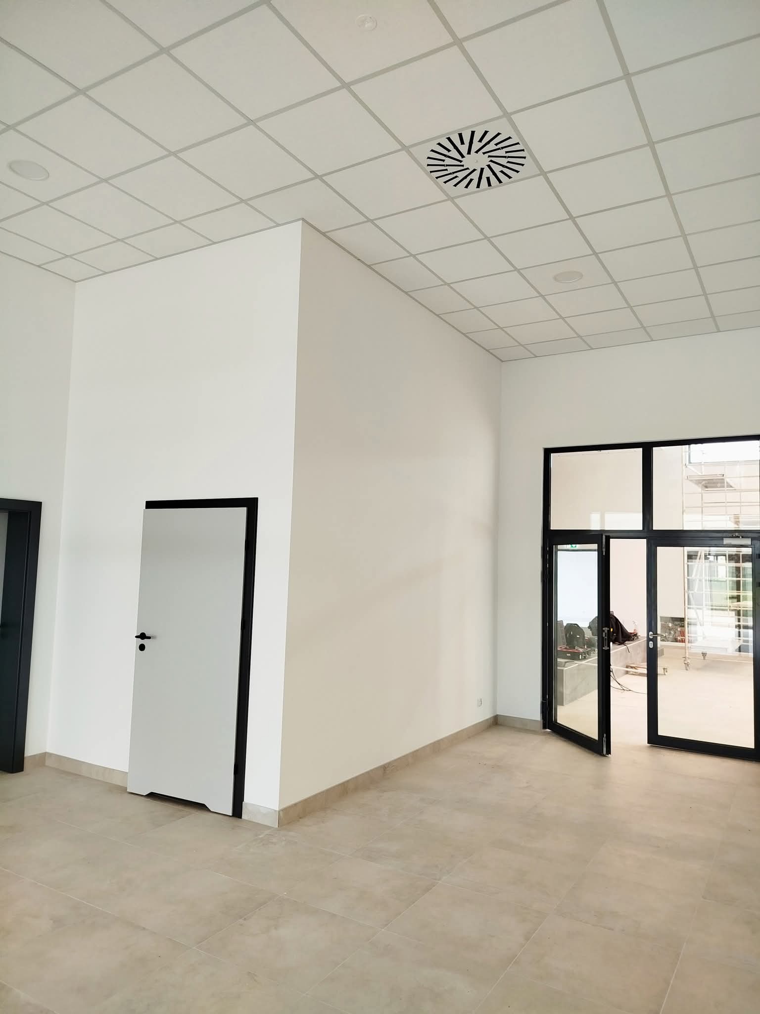 Jasne wnętrze biurowe z białymi ścianami, płytkami podłogowymi i sufitem kasetonowym. Czarne ramy drzwi i okien kontrastują z bielą ścian. Widoczne elementy instalacji sufitowej.