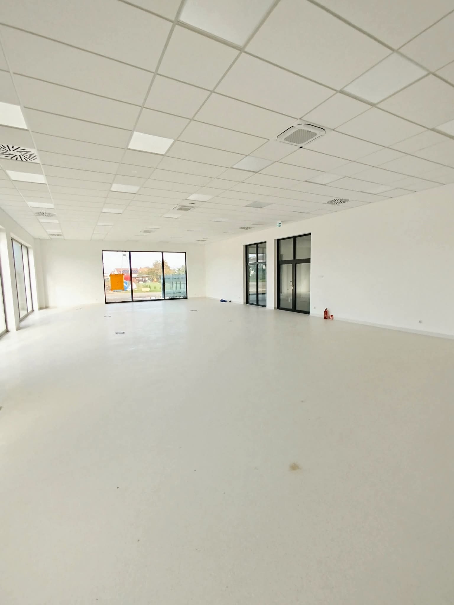 Jasne, przestronne wnętrze biurowe z dużymi oknami i białymi ścianami. Widoczne panele sufitowe, wentylacja i drzwi. Minimalistyczny design, świeżo po wykończeniu.