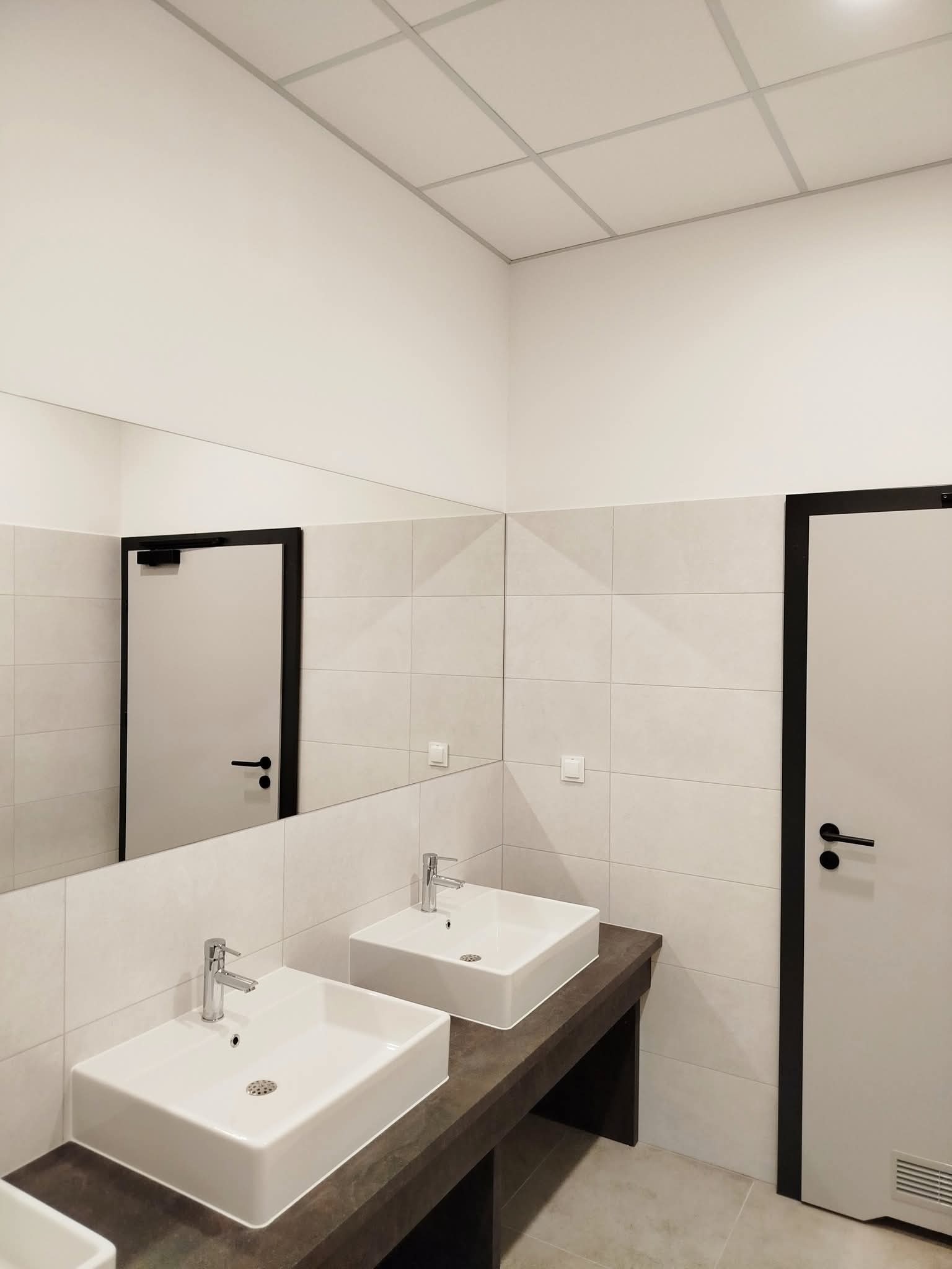 Minimalistyczna łazienka z dwoma umywalkami nablatowymi, jasnymi płytkami i czarnymi akcentami w postaci ramy drzwi i klamek. Nowoczesne wykończenie.
