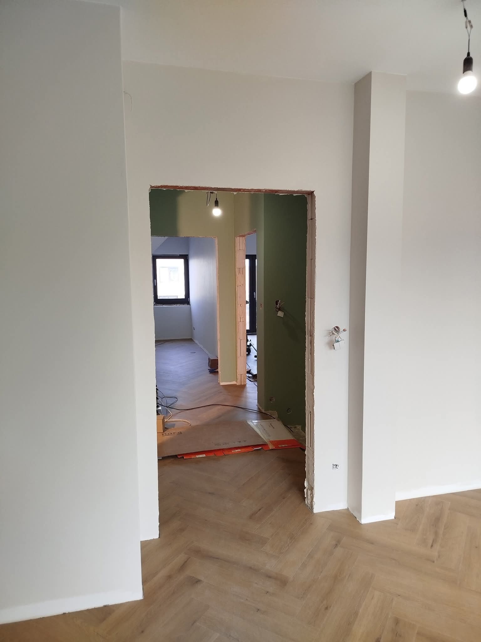 Świeżo pomalowane, jasne ściany w pomieszczeniu z parkietem ułożonym w jodełkę, widok na sąsiednie pokoje przez otwór drzwiowy w trakcie prac wykończeniowych.