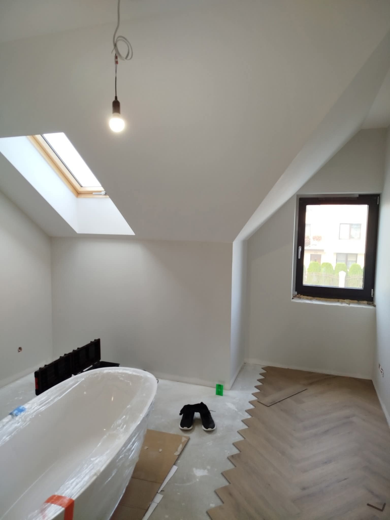 Poddasze w trakcie wykańczania: białe ściany, okno dachowe, ułożona część parkietu w jodełkę, wanna zabezpieczona folią. Widoczna żarówka na kablu.