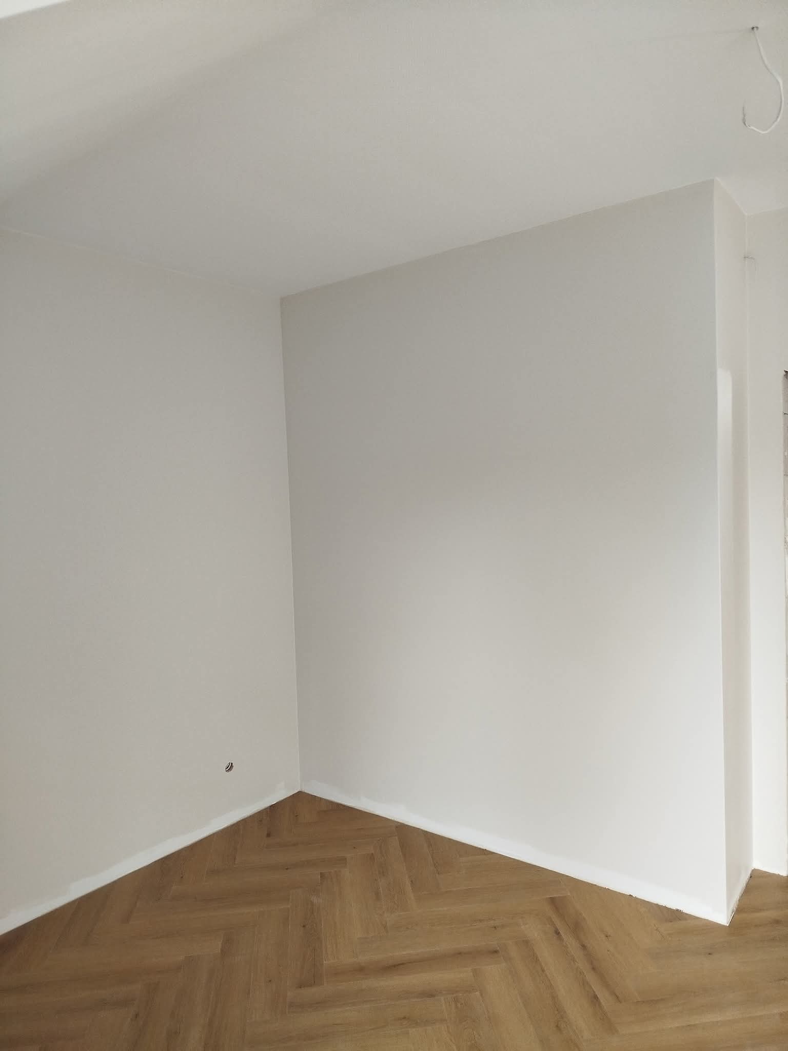 Jasne wnętrze po remoncie: świeżo pomalowane ściany w odcieniach bieli i parkiet ułożony w jodełkę. Widoczne gniazdko elektryczne i wystający kabel ze ściany.