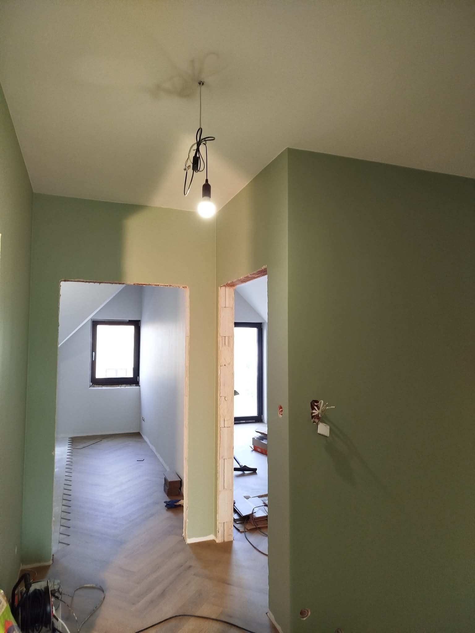 Wnętrze w trakcie prac wykończeniowych: świeżo pomalowane ściany w odcieniach zieleni, parkiet ułożony w jodełkę, widoczne instalacje elektryczne i przygotowania do montażu ościeżnic.