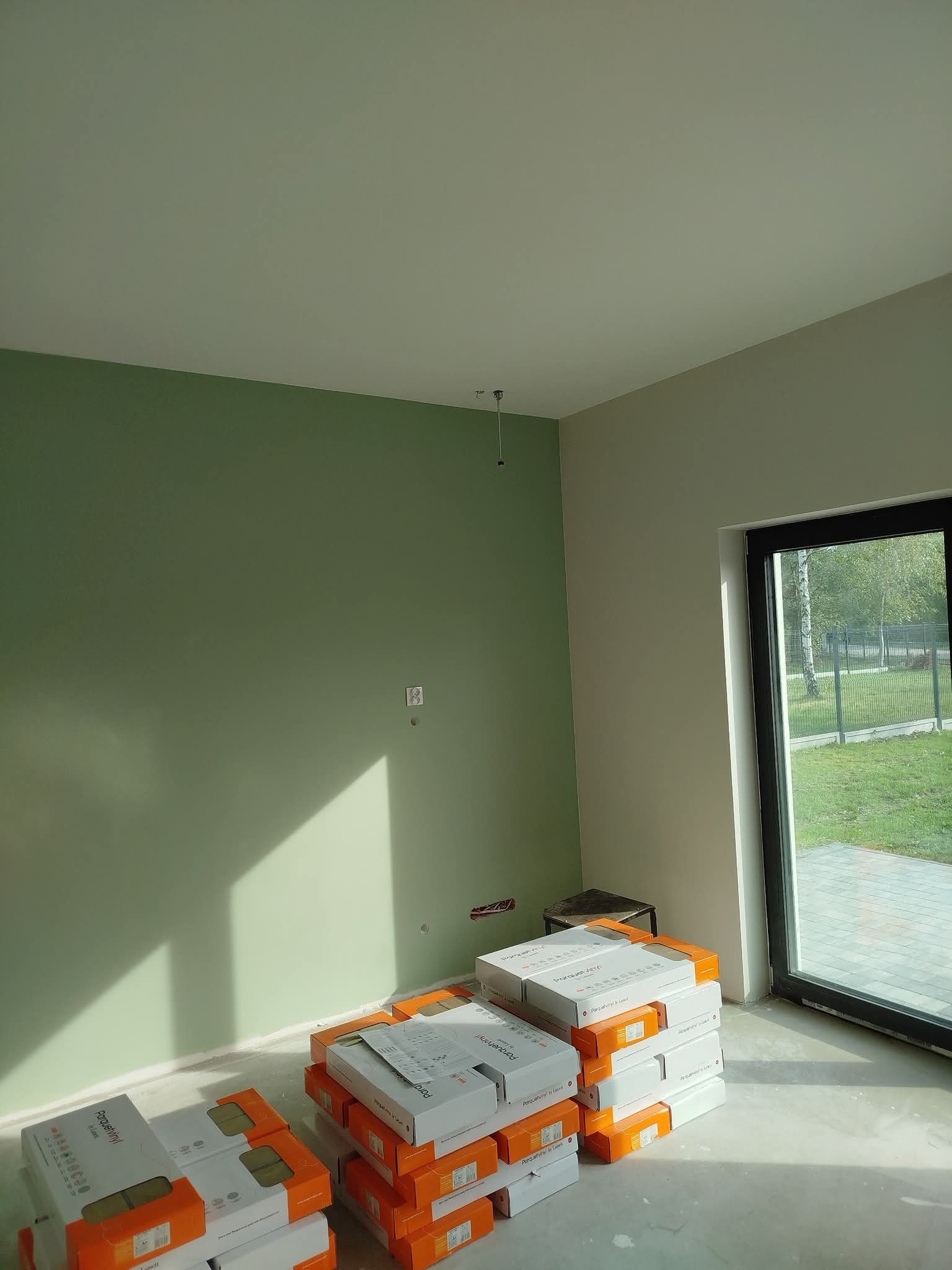 Świeżo pomalowane na zielono ściany w pokoju z oknem. Na podłodze stosy paneli gotowych do ułożenia. Widoczne przygotowania do prac wykończeniowych.