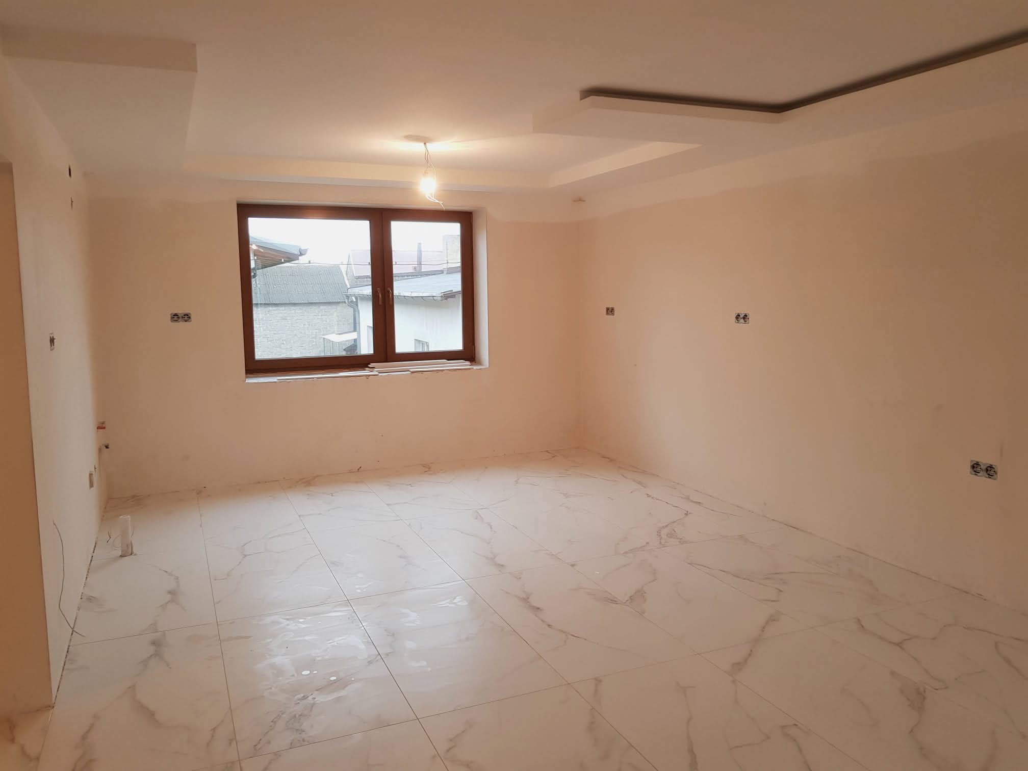 Jasne wnętrze po remoncie: marmurowe płytki podłogowe, gładkie, beżowe ściany, okno z drewnianą ramą i ozdobny sufit podwieszany z centralnym punktem świetlnym.