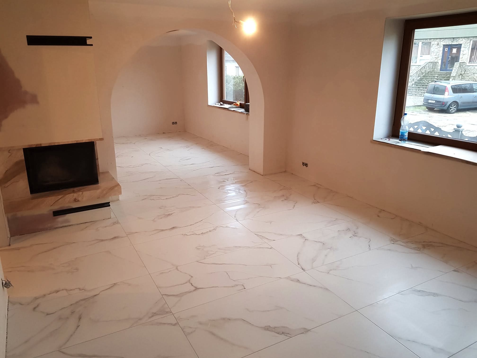 Jasne płytki imitujące marmur na podłodze przestronnego wnętrza z kominkiem i łukowym przejściem. Ściany w trakcie prac wykończeniowych, widoczne gniazdka.