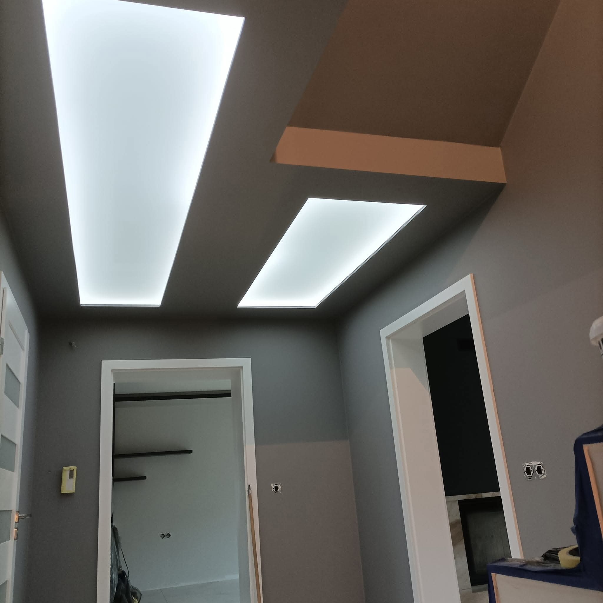 Szare wnętrze z oświetleniem sufitowym LED w kształcie prostokątów, widoczne drzwi z białymi ościeżnicami i fragment ściany. Minimalistyczny design.