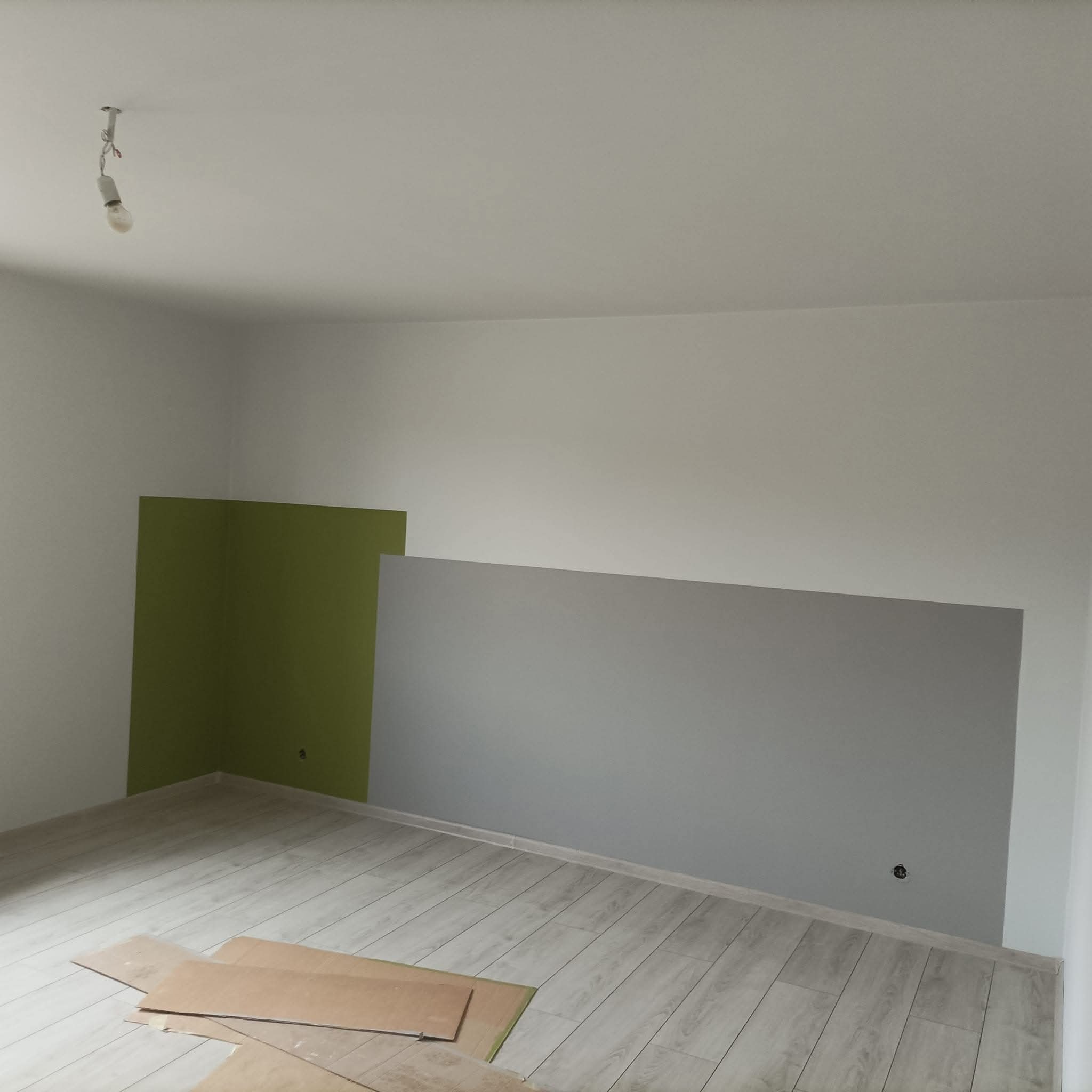 Świeżo pomalowane ściany w pokoju, fragmenty w kolorze zielonym i szarym, podłoga wyłożona panelami. Na podłodze leżą kartony zabezpieczające.