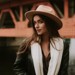 Portret młodej kobiety w kapeluszu i skórzanej kurtce, z białym szalem, na tle przęsła mostu w pochmurny dzień.