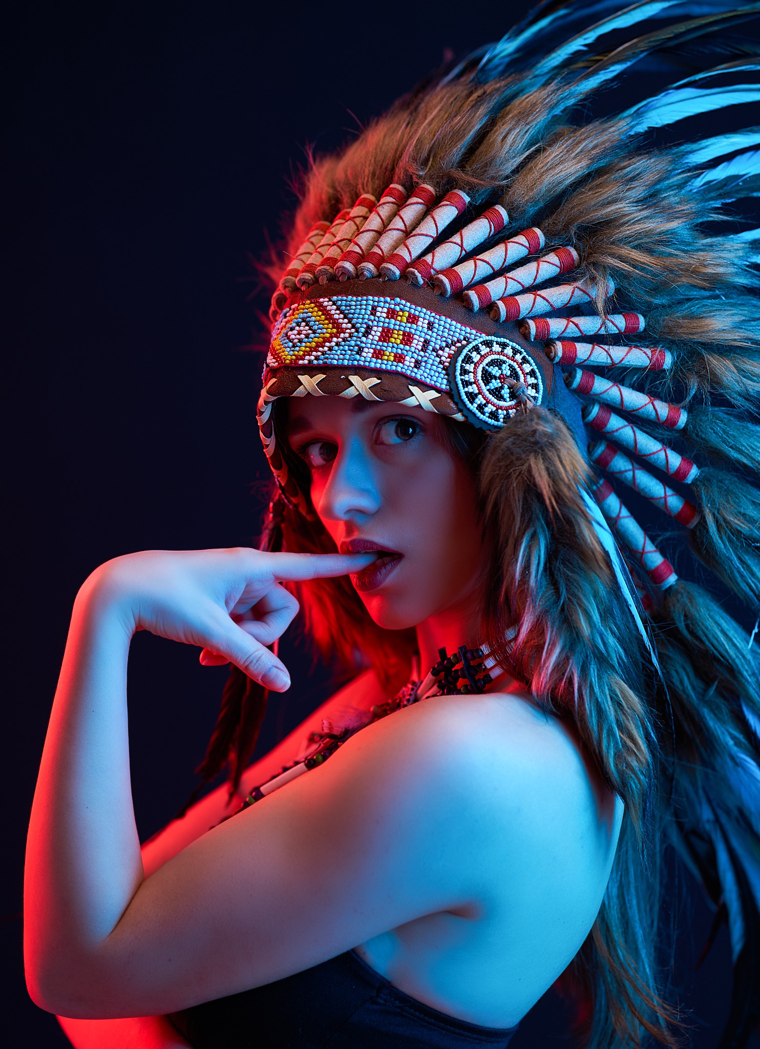 Portret kobiety w nakryciu głowy z piór w stylu indiańskim, oświetlonej czerwonym i niebieskim światłem, z palcem przy ustach.