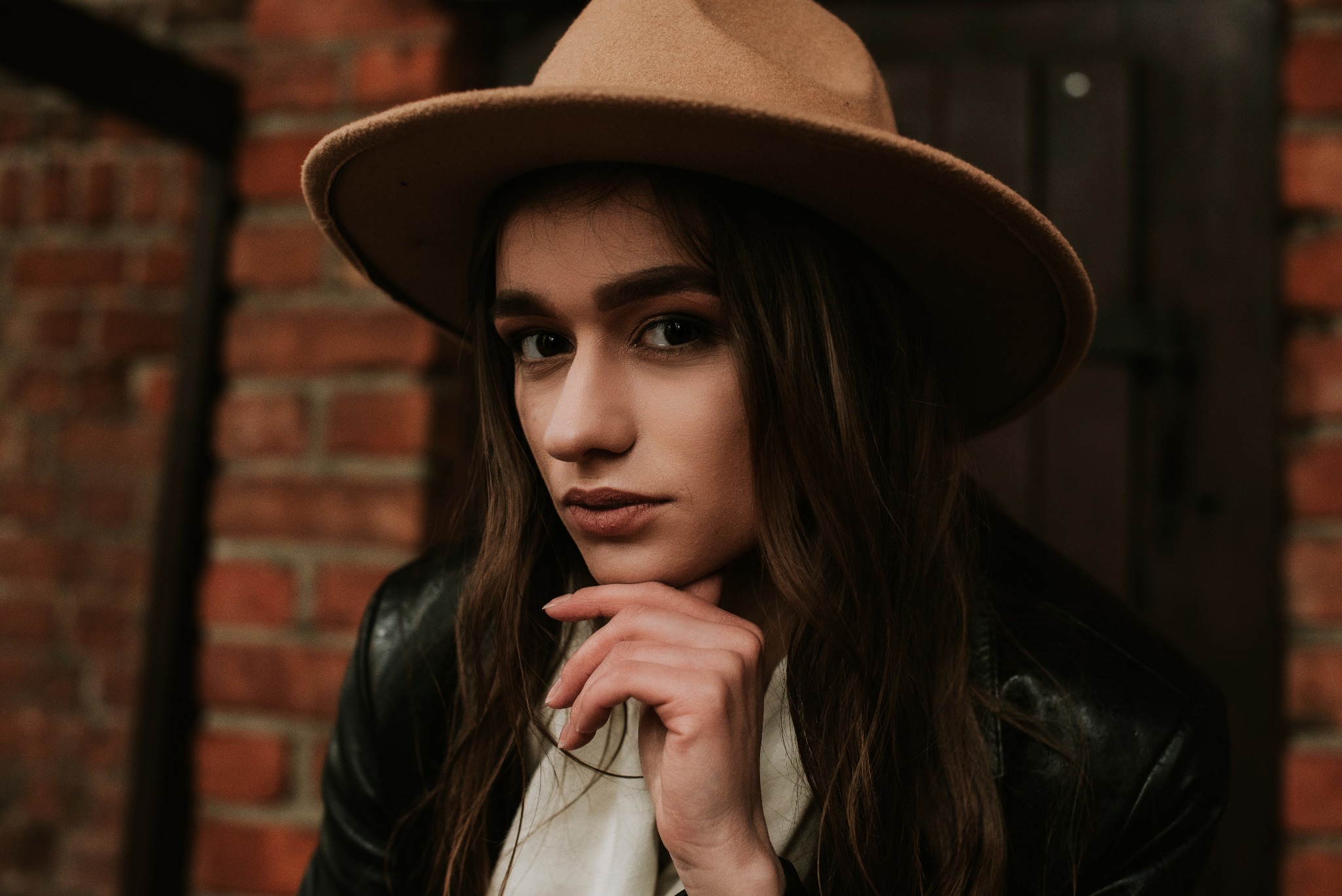 Portret młodej kobiety w brązowym kapeluszu, skórzanej kurtce i białej bluzce, opierającej podbródek na dłoni na tle ceglanej ściany.