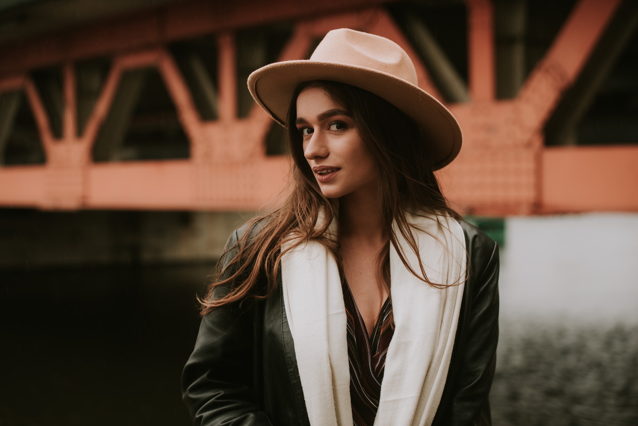 Portret młodej kobiety w kapeluszu i skórzanej kurtce, z białym szalem, na tle przęsła mostu w pochmurny dzień.