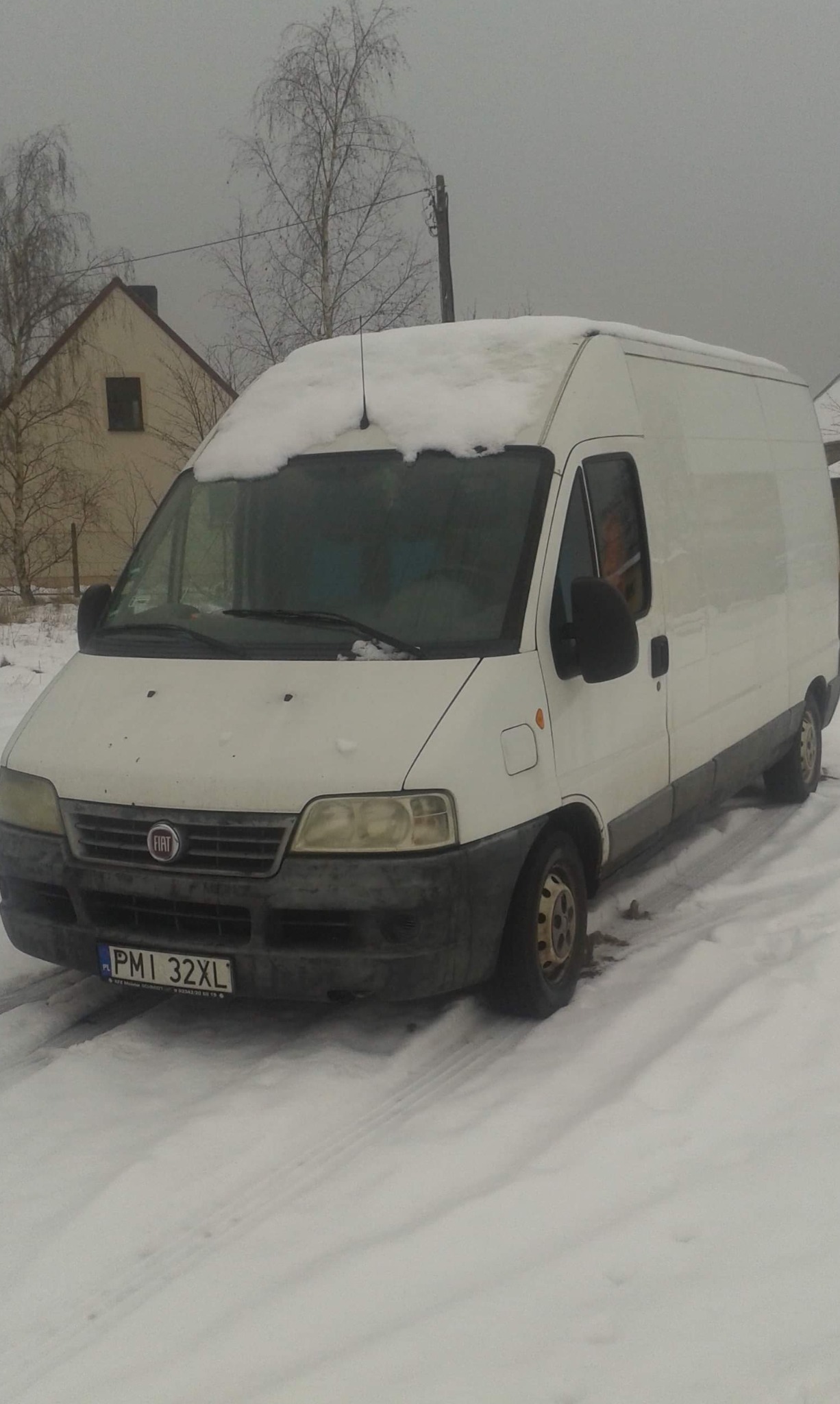 Biały Fiat Ducato zaparkowany na śniegu przed domem w pochmurny dzień, z lekką warstwą śniegu na dachu.