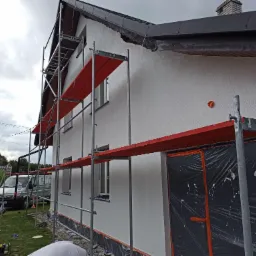 Dom jednorodzinny w trakcie ocieplania elewacji, widoczne rusztowanie z czerwonymi deskami, zabezpieczone okna i drzwi folią ochronną, świeżo nałożony jasny tynk.