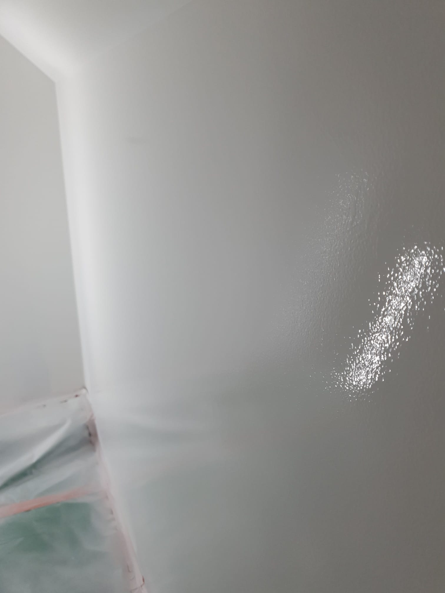 Świeżo pomalowana, jasnoszara ściana w rogu pomieszczenia, z widocznym zabezpieczeniem podłogi folią malarską.