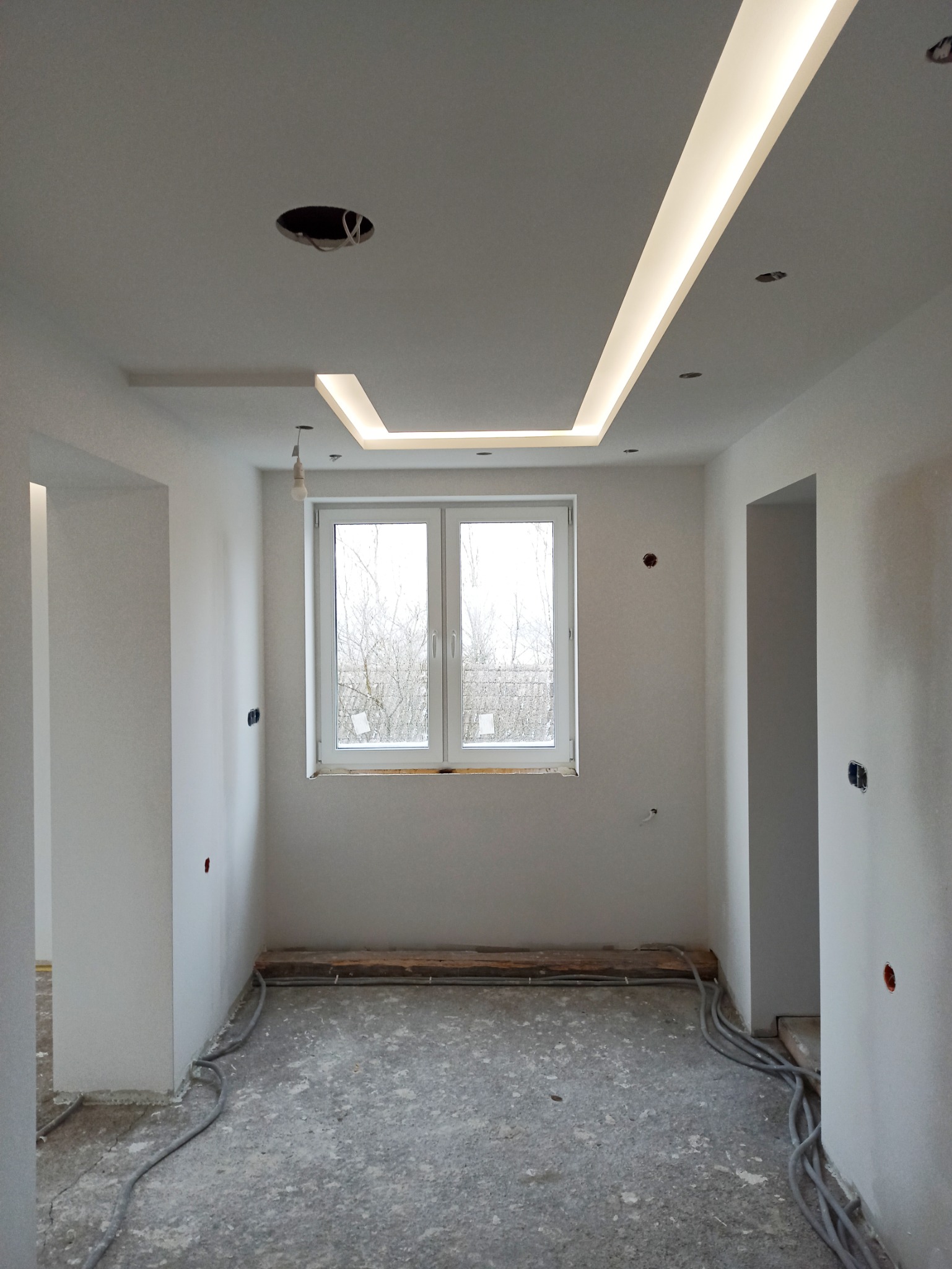 Pokój w trakcie prac wykończeniowych: białe ściany, surowa podłoga, okno z widokiem na drzewa, podwieszany sufit z oświetleniem LED, widoczne kable elektryczne.