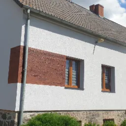 Otynkowana na biało ściana domu z dekoracyjnymi, brązowymi panelami imitującymi drewno, widoczne okna z roletami, kamienny fundament i fragment dachu oraz osoba w roboczym stroju.