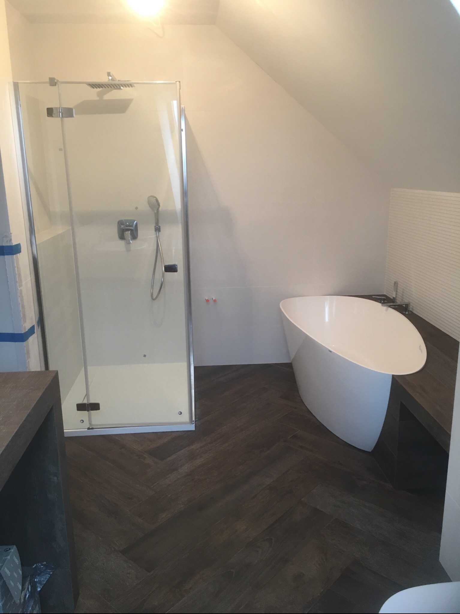 Nowoczesna łazienka na poddaszu ze szklaną kabiną prysznicową, wolnostojącą wanną i parkietem ułożonym w jodełkę.