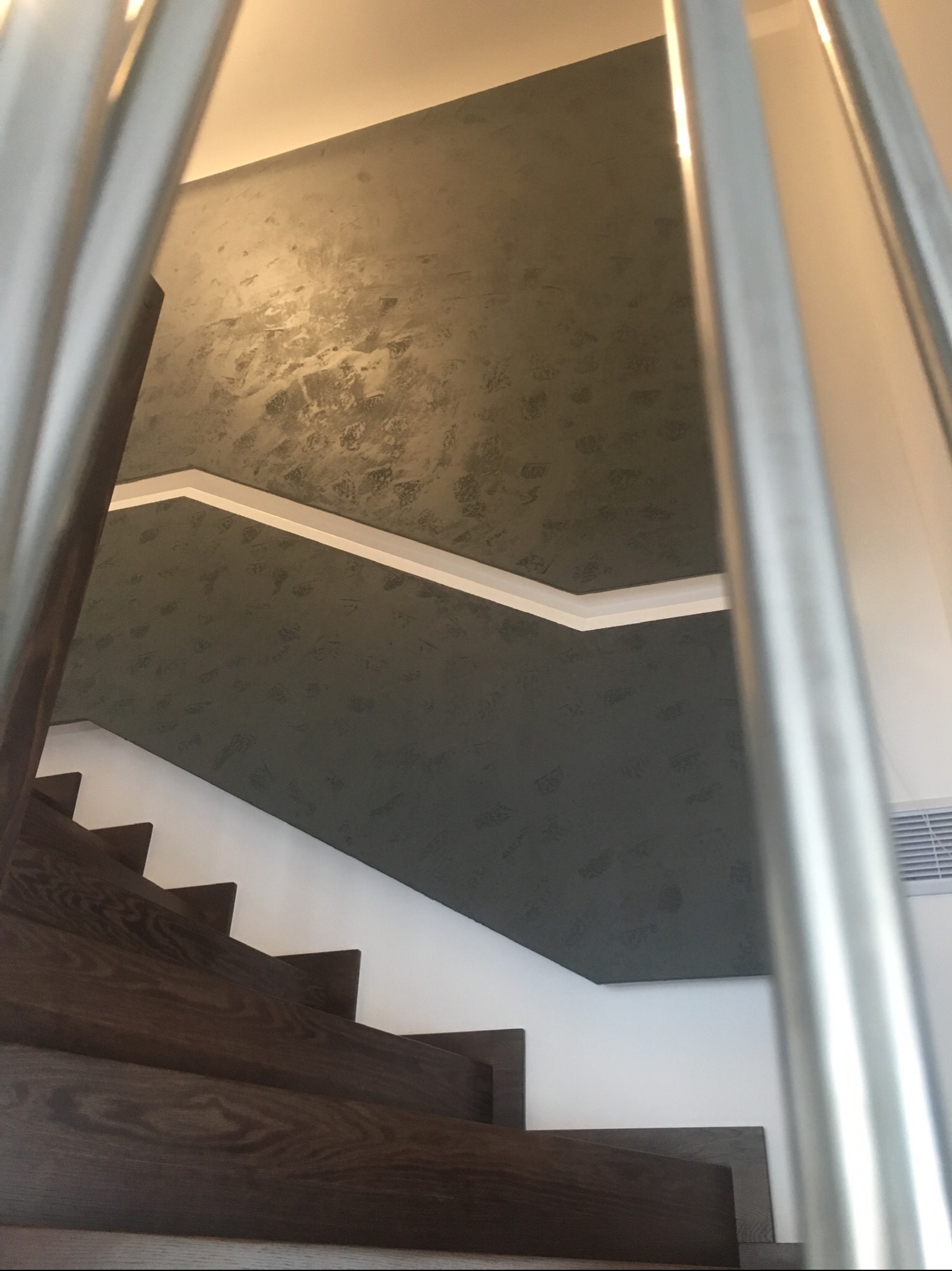 Wnętrze klatki schodowej z drewnianymi schodami i ścianą pokrytą dekoracyjną szarozieloną farbą z efektem cieniowania, widok przesłonięty przez metalowe barierki.