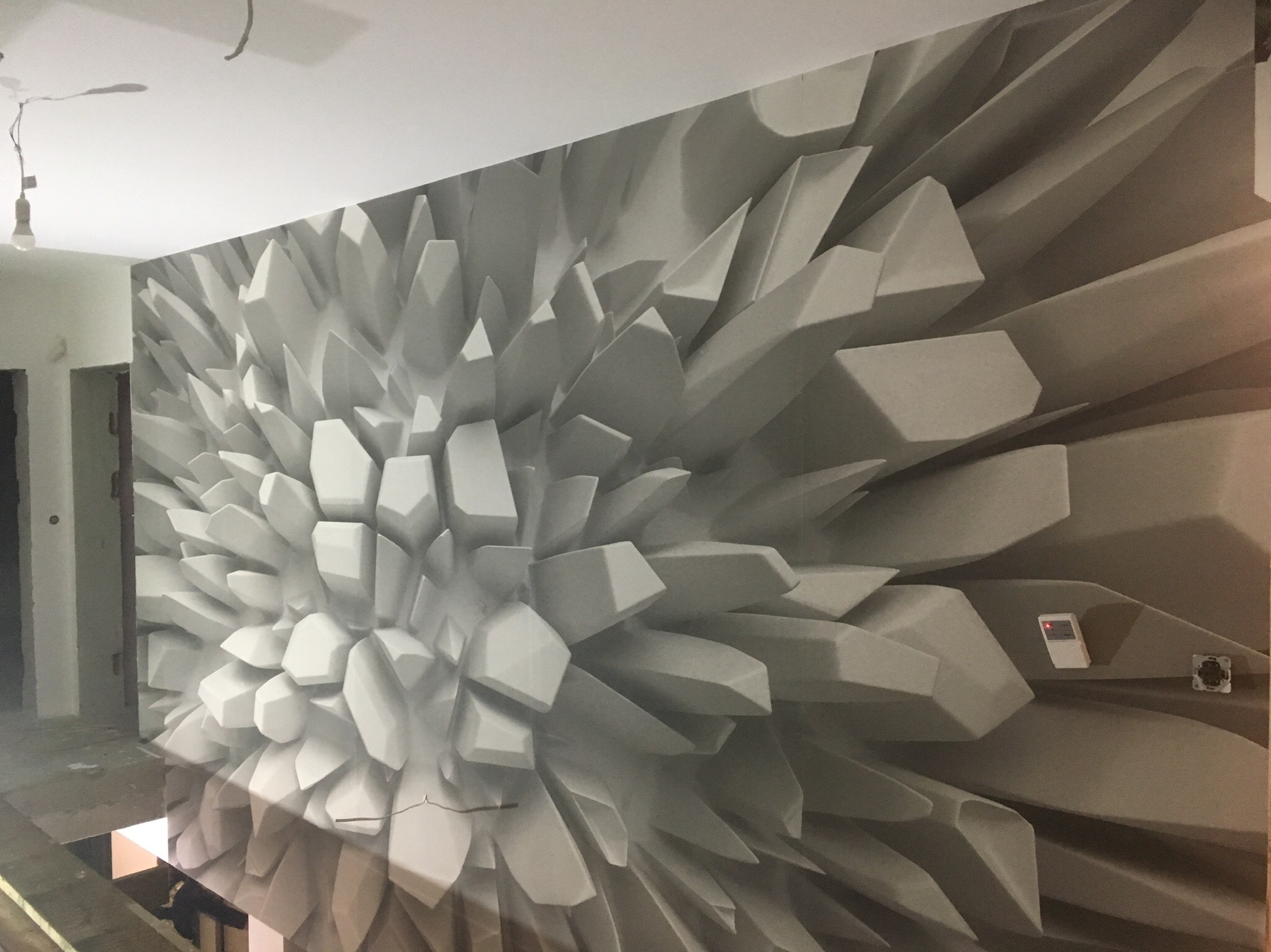 Fototapeta 3D z efektem geometrycznym w odcieniach szarości, imitująca rozkwitający kryształ, z widocznym panelem sterowania i gniazdkiem elektrycznym na ścianie.