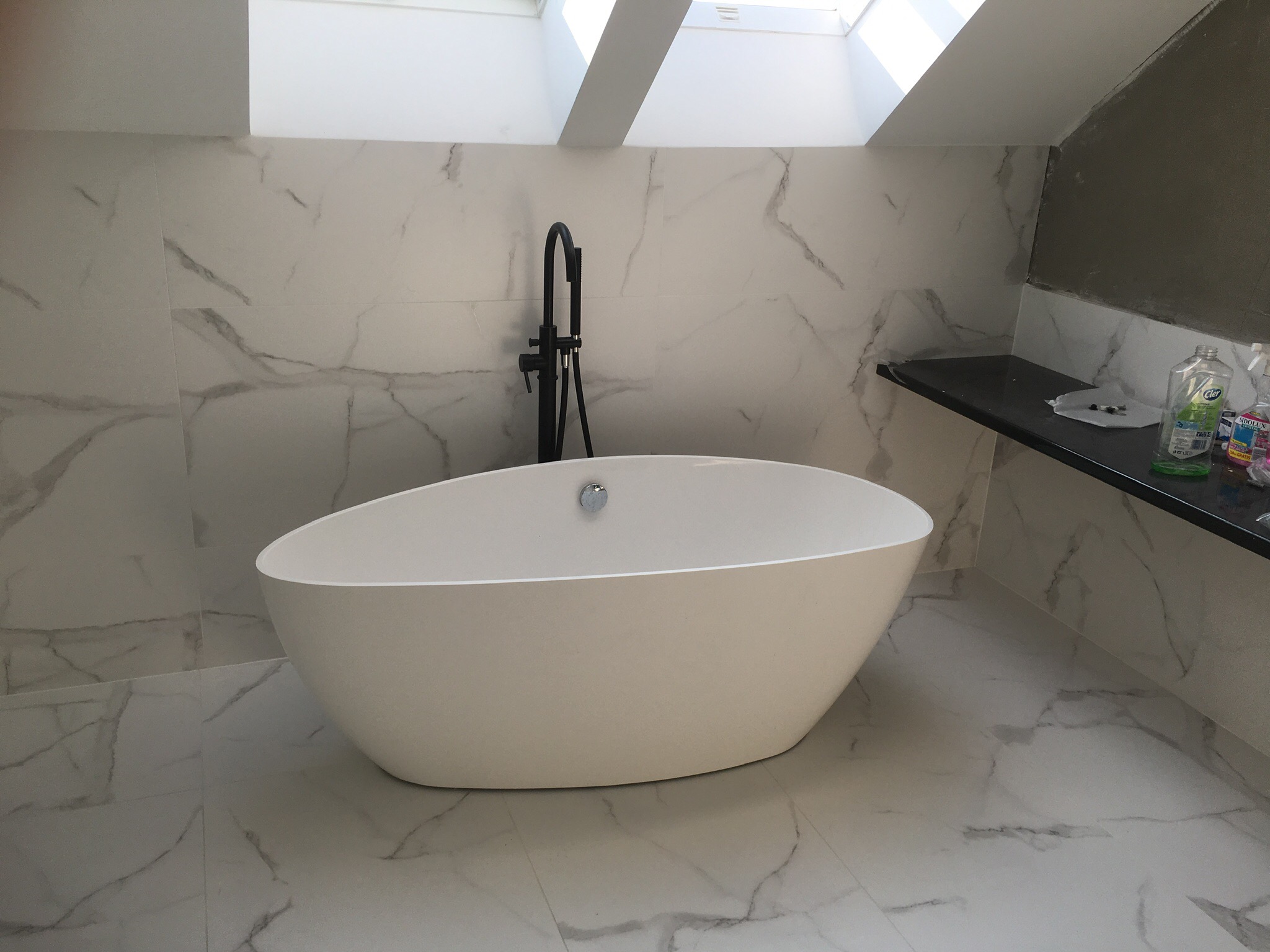 Nowoczesna, wolnostojąca wanna owalna z czarną armaturą na tle marmurowych płytek w łazience z oknem dachowym.