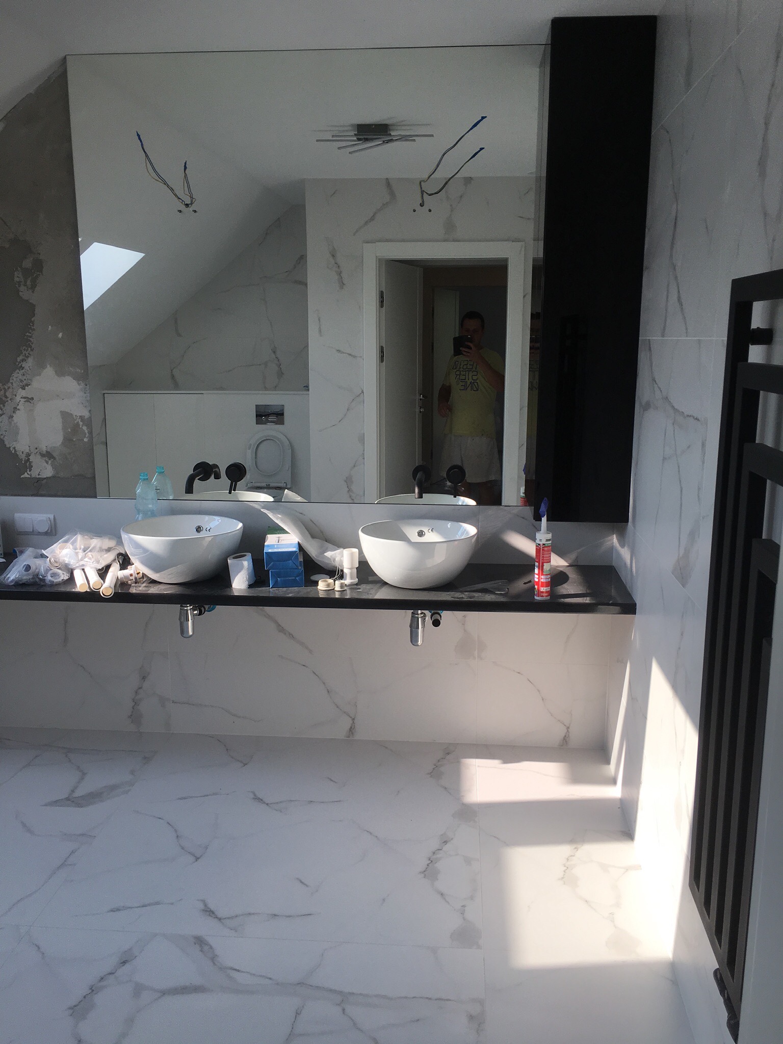 Łazienka w trakcie montażu: dwie białe umywalki nablatowe, czarne baterie, marmurowe płytki na ścianach i podłodze, widoczne narzędzia i materiały instalacyjne, w lustrze odbija się osoba robiąca...