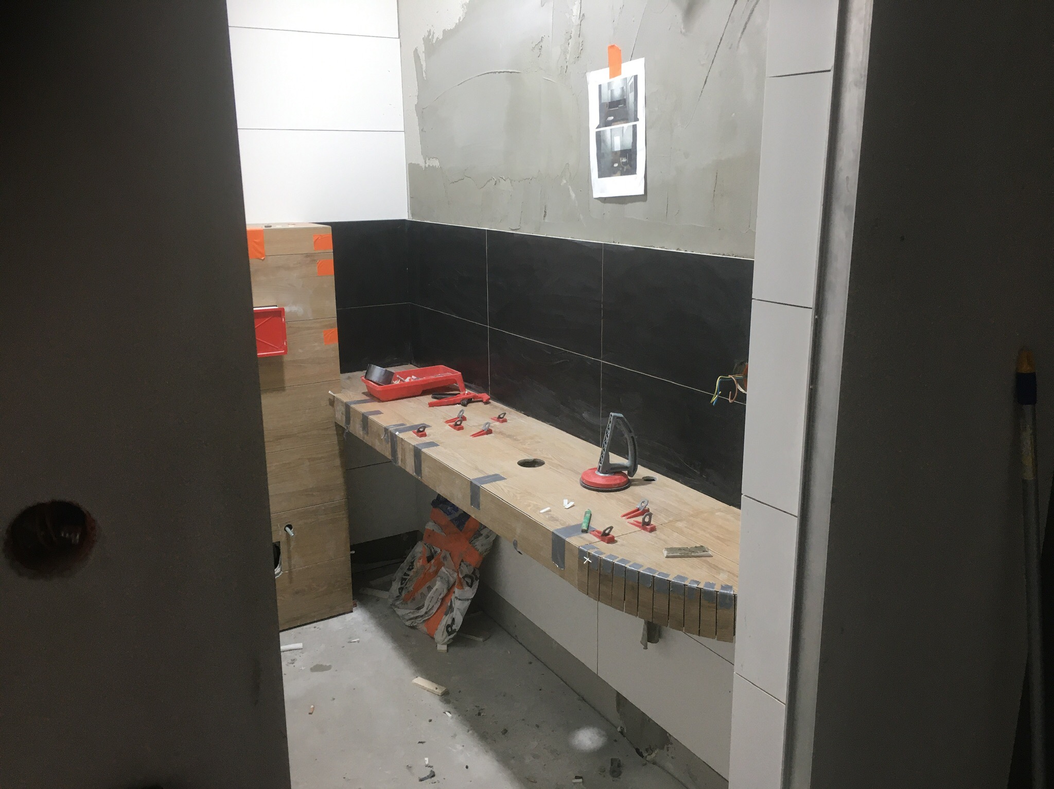 Montaż blatu w łazience z użyciem przyssawek i klipsów poziomujących, widoczne czarne i białe płytki na ścianie, remont w trakcie.