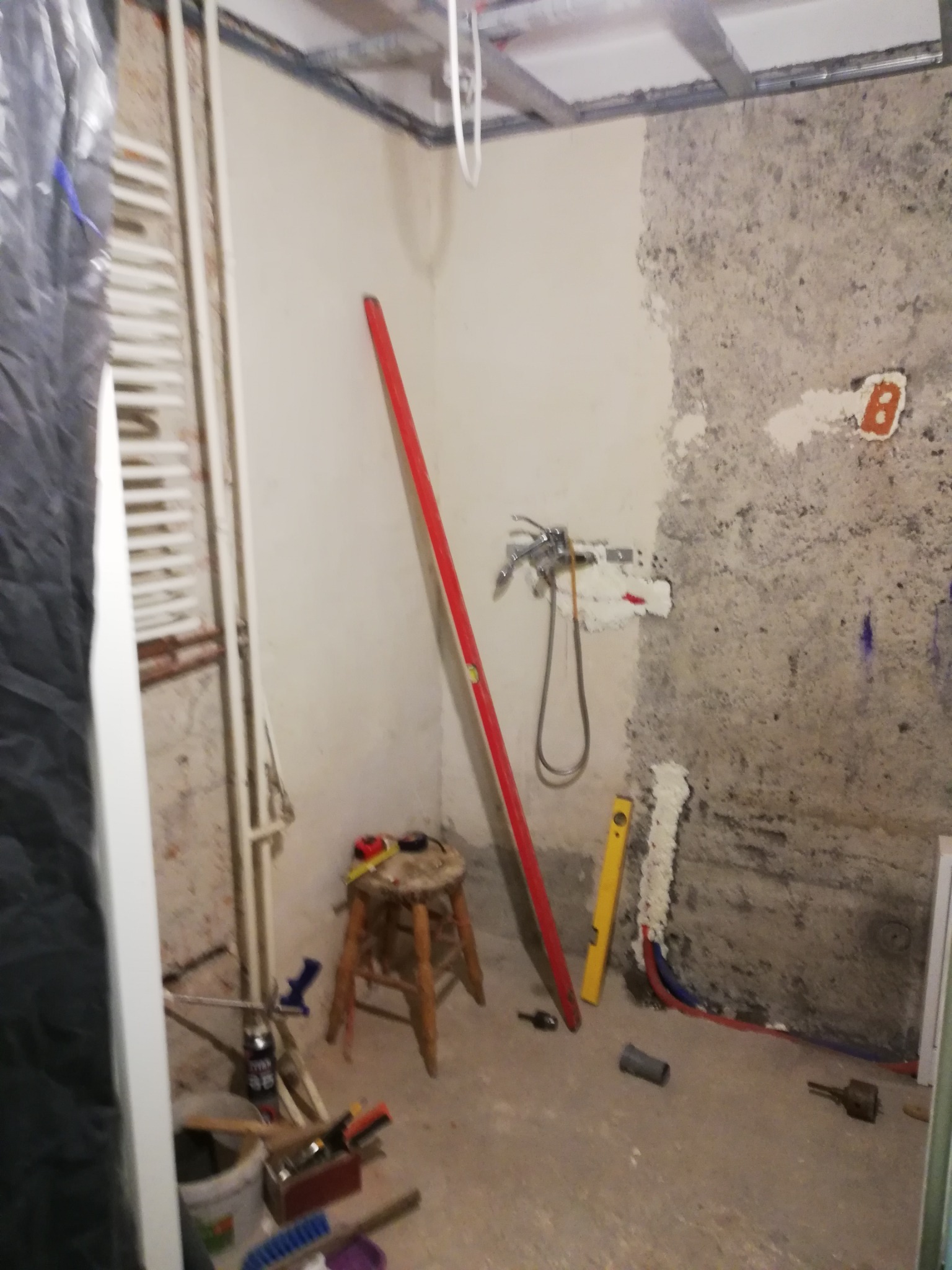 Łazienka w trakcie remontu: częściowo otynkowana ściana z zamontowaną baterią, pionowa czerwona poziomica oparta o ścianę, narzędzia na podłodze i stołku, grzejnik drabinkowy po lewej stronie.