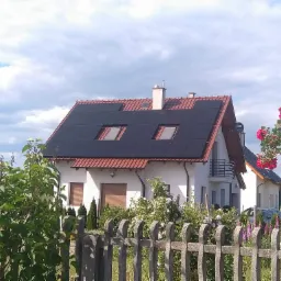 Dom jednorodzinny z zamontowanymi panelami słonecznymi na dachu, widok zza drewnianego płotu i kwitnących krzewów.