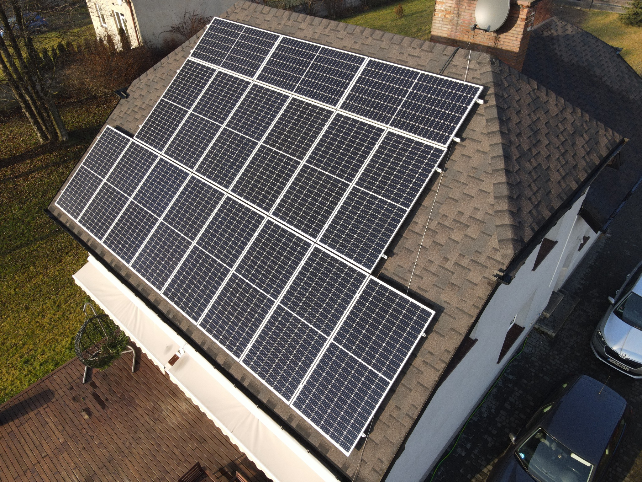 Widok z góry na panele słoneczne zamontowane na dachu domu z brązową dachówką bitumiczną, obok komin z anteną satelitarną, na dole taras drewniany i dwa samochody na podjeździe.