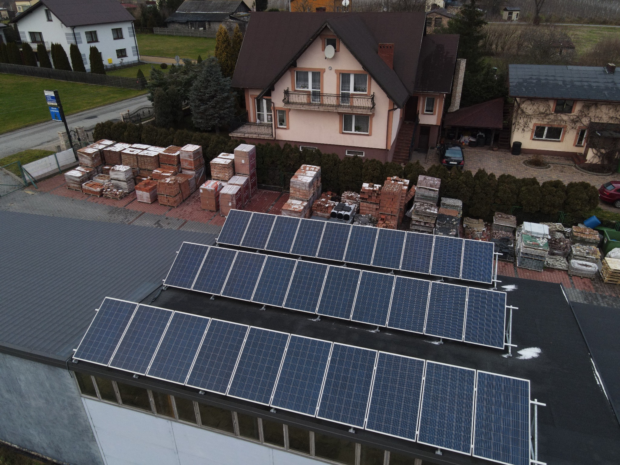 Widok z góry na rzędy paneli słonecznych na dachu budynku, z paletami cegieł w tle i domami w oddali.