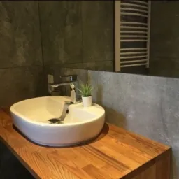 Nowoczesna łazienka z umywalką nablatową na drewnianym blacie, szare płytki na ścianach i grzejnik drabinkowy.