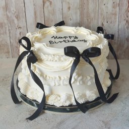 Sama Słodycz - Dwupiętrowy tort urodzinowy z białym kremem i czarnymi wstążkami na drewnianym tle.