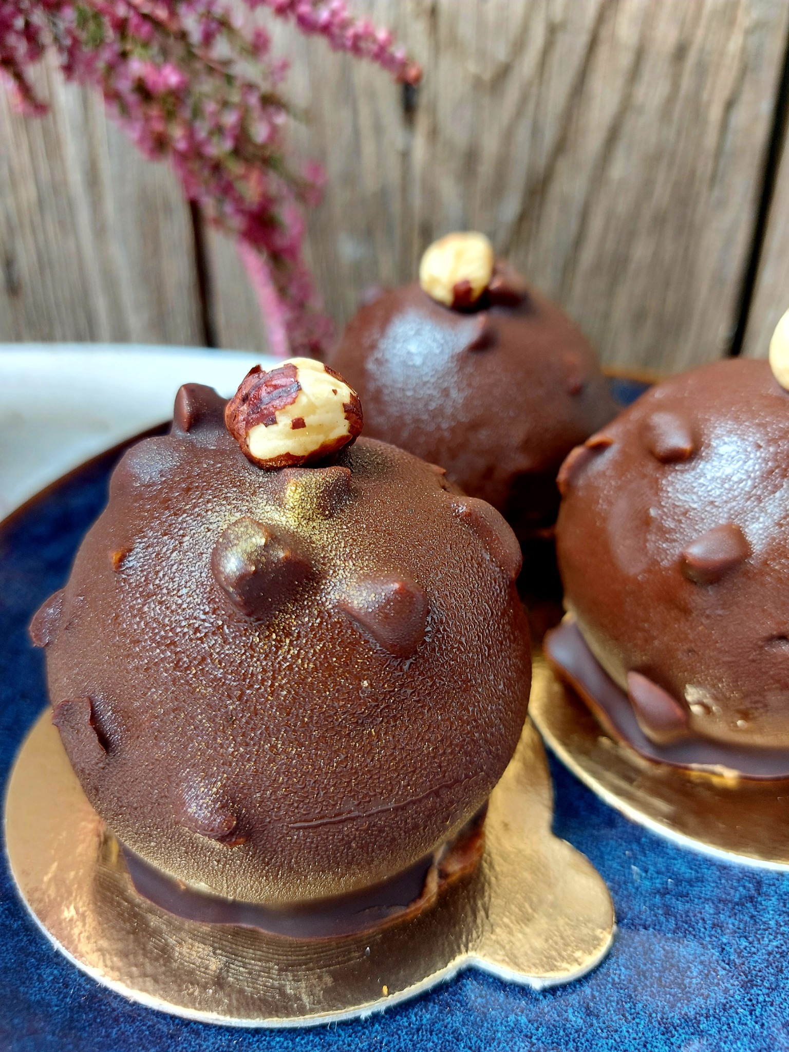 Trzy czekoladowe kulki deserowe z orzechami laskowymi na niebieskim talerzu z kwiatami wrzosu w tle.