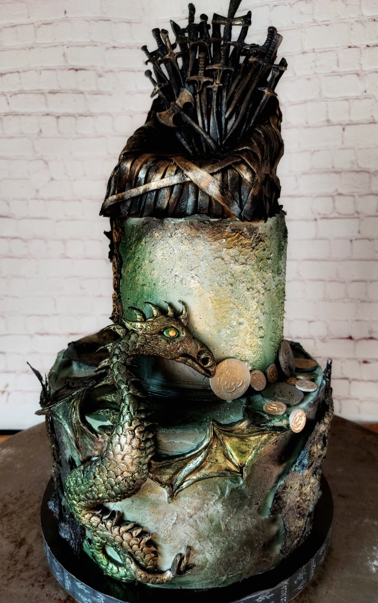 Trzypiętrowy tort zdobiony motywami z Gry o Tron: Żelazny Tron na szczycie, zielony smok owinięty wokół dolnej warstwy, złote monety na środkowym poziomie, tło z białej cegły.