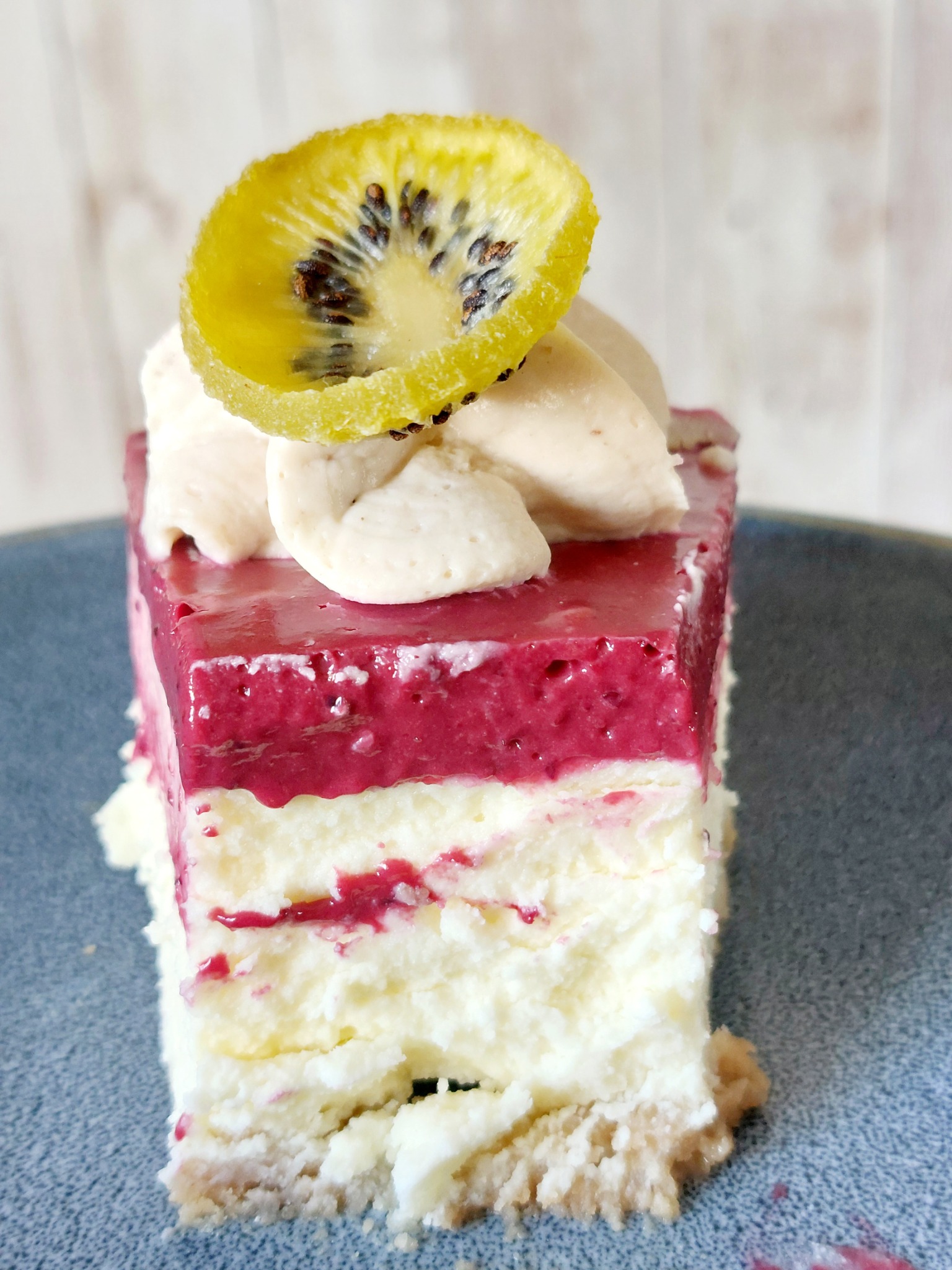 Kwadratowy kawałek ciasta z kremem i galaretką malinową, udekorowany plasterkiem złotego kiwi na niebieskim talerzu.