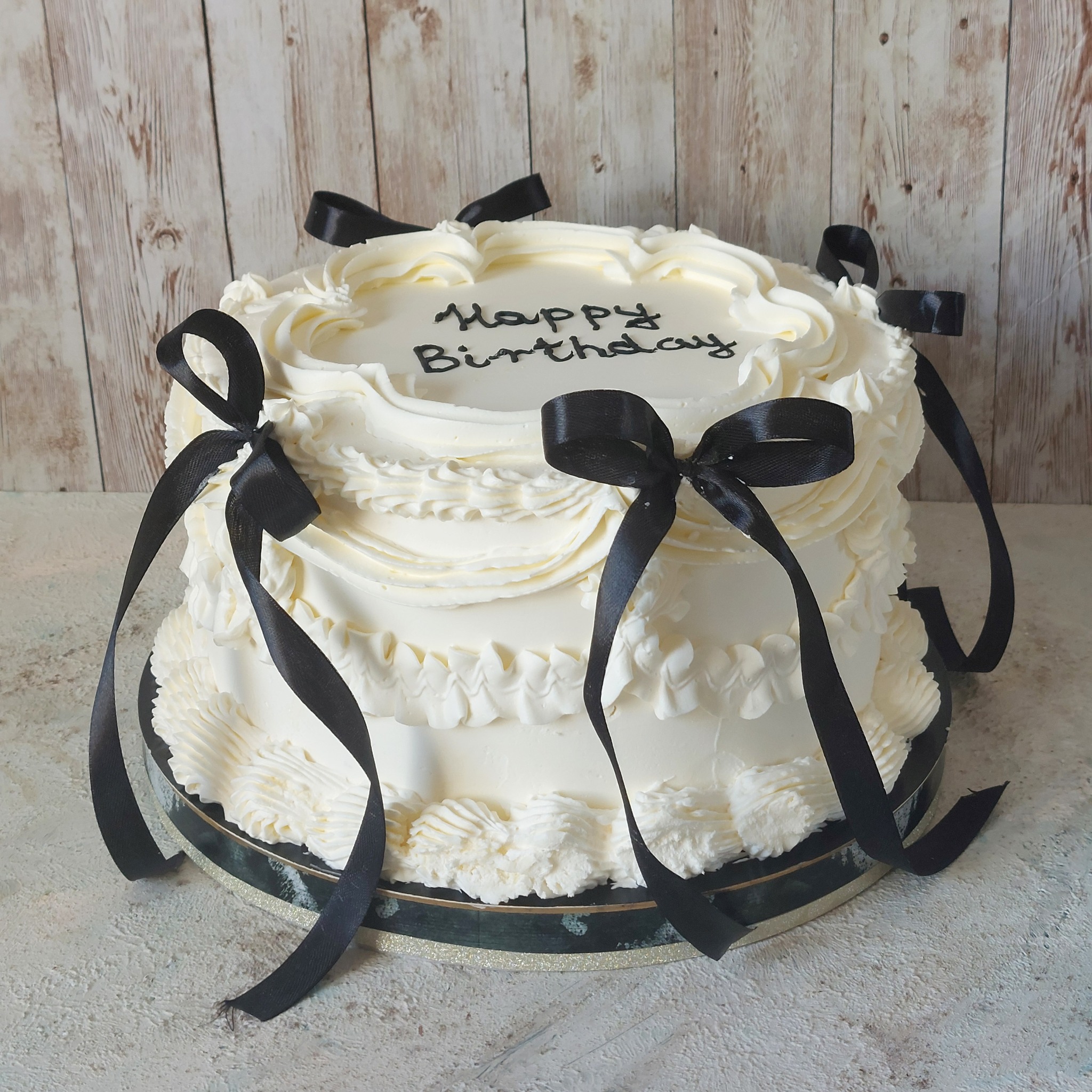 Dwupiętrowy tort urodzinowy z białym kremem i czarnymi wstążkami na drewnianym tle.