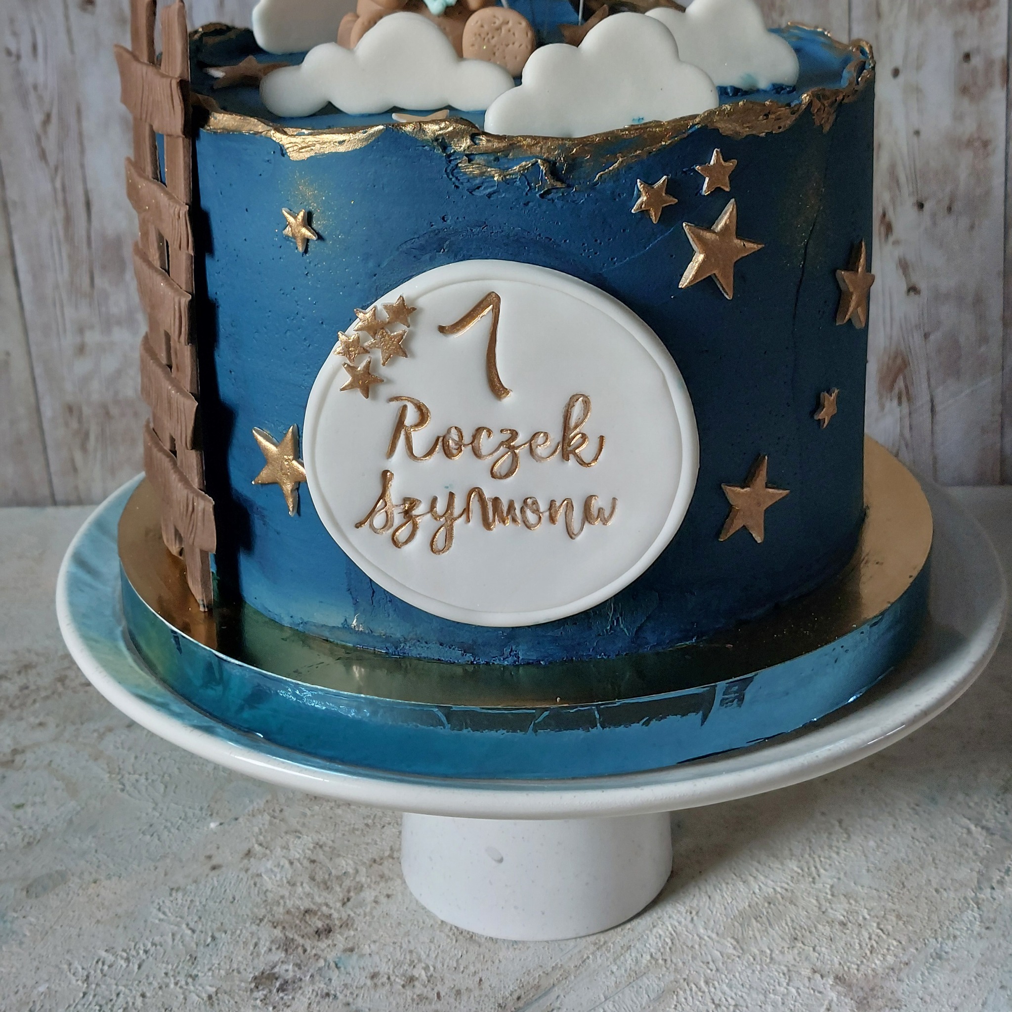 Tort na pierwsze urodziny Szymona, zdobiony motywem nocnego nieba z gwiazdami i chmurkami, ustawiony na niebieskim paterze.