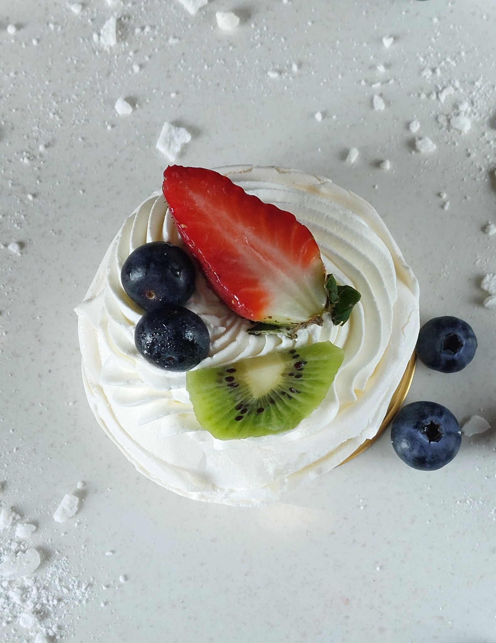 Mini beza z kremem, truskawką, kiwi i borówkami na białym tle z drobnymi okruchami.