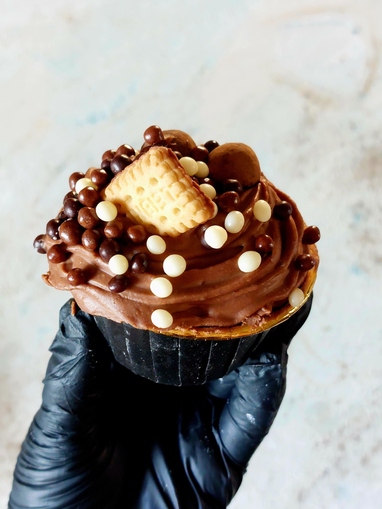 Czekoladowy cupcake z kremem, posypką z kulek i herbatnikiem, trzymany w dłoni w czarnej rękawiczce.