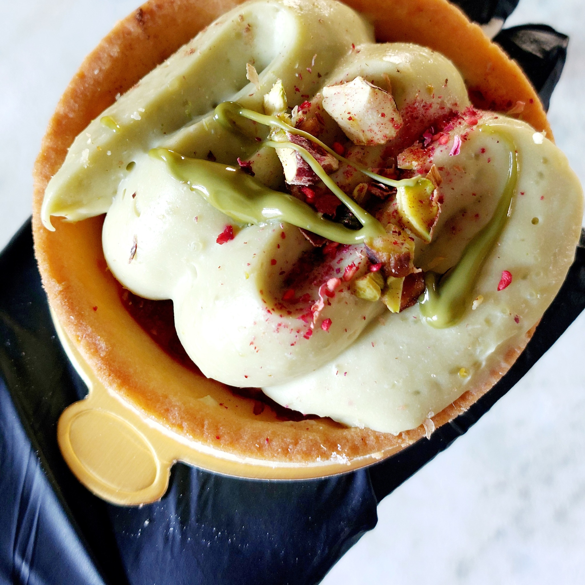 Tarta z kremem pistacjowym, posypana liofilizowanymi malinami i pistacjami, na dłoni w czarnej rękawiczce.